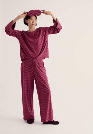 Personne portant un pyjama et des chaussettes rayés bordeaux assortis, tenant un masque de sommeil rayé bordeaux au-dessus de la tête.