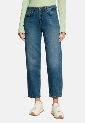 Femme portant un jean bleu taille haute à jambes droites, un haut à manches longues côtelé vert clair, et des baskets épaisses blanc cassé.