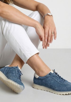 Person trägt weiße Hose und blaue Veloursleder-Sneaker, sitzt mit den Händen auf dem gekreuzten Knie, trägt eine Uhr und Ringe.