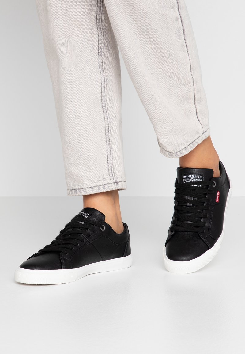 Sneakers in pelle nera con suole e lacci in gomma bianca. Presentano un piccolo logo rosso sul lato e una superficie liscia. Indossati con pantaloni chiari.