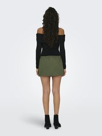 Žena stojí chrbtom k nám, má na sebe čierny top s odhalenými ramenami, olivovo zelenú minisukňu a čierne členkové topánky na jednoliatom svetlom pozadí.