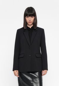 Donna che indossa un blazer nero su una blusa nera e una gonna di pelle nera, in piedi contro uno sfondo bianco semplice.