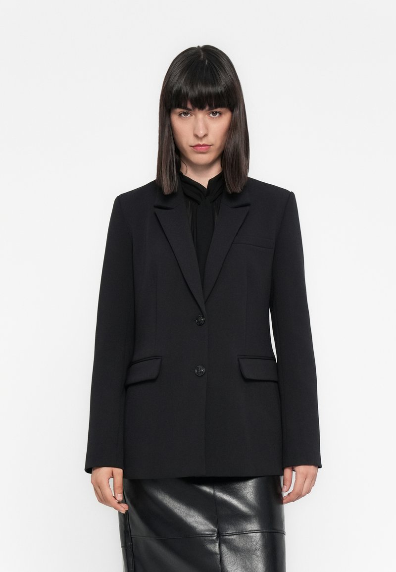 Donna che indossa un blazer nero su una blusa nera e una gonna di pelle nera, in piedi contro uno sfondo bianco semplice.