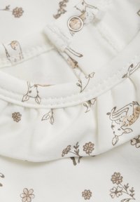 Vitt tyg med bruna blommönster och hjortmönster. Har en volangkrage och tryckknapp för enkel klädsel. Mjukt material.