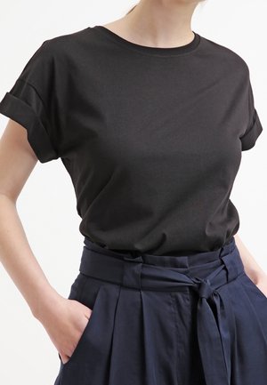 Kobieta w dopasowanym czarnym, krótkim rękawem t-shircie wpuszczonym w granatowe spodnie z wysokim stanem, plisowane, z paskiem w talii, ręce w kieszeniach.