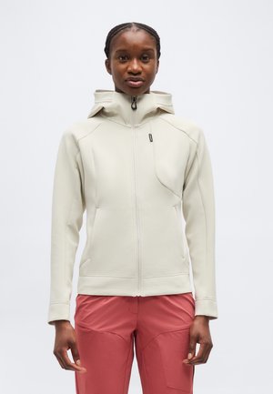 Nő világos bézs cipzáras kapucnis kabátban és korallszínű rózsaszín nadrágban, előre nézve, karjai oldalt lazán lelógva.