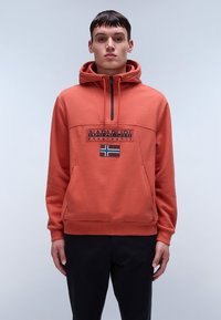 Felpa arancione con cappuccio e collo con zip, caratterizzata da una patch logo nera e un piccolo design a bandiera. Realizzata in materiale morbido e testurizzato.