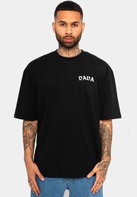 Schwarzes Baumwoll-T-Shirt mit kurzen Ärmeln, lockerer Passform, mit weißem "DADA"-Logo auf der linken Brust. Getragen mit hellblauen Jeans.