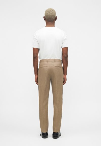 Άνδρας με κοντά ξανθά μαλλιά στέκεται με την πλάτη, φορώντας λευκό κοντομάνικο πουκάμισο, μπεζ παντελόνι και μαύρα παπούτσια σε λευκό φόντο.