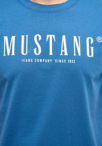 T-shirt bleu avec le texte blanc "MUSTANG" et un texte plus petit "JEANS COMPANY SINCE 1932" centré sur la poitrine.