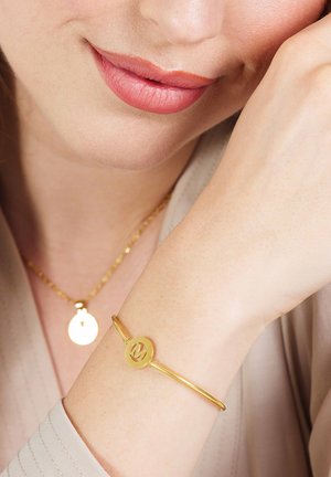 Goldene Metallarmband mit einem runden Anhänger, der ein Buchstabendesign zeigt. Wird am Handgelenk getragen und mit einer passenden Halskette kombiniert, beide schlicht im Stil.