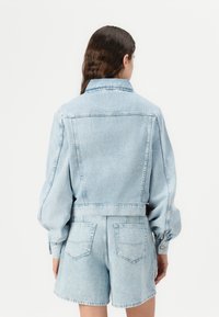 Veste en denim bleu clair au design court, avec des poignets à boutons et des poches latérales, accompagnée de shorts en denim assortis. Surface texturée visible.