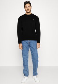 Čierny pletený sveter s dlhými rukávmi, okrúhlym výstrihom a malým vyšívaným logom. Kombinovaný so svetlo modrými rifľami a bielymi teniskami.