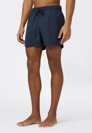 Hombre con pantalones cortos de baño azul oscuro con cintura elástica y cordón, de pie descalzo contra un fondo liso.