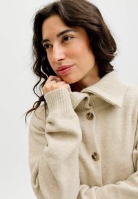 Donna con capelli scuri e mossi, che indossa un maglione a maglia beige con bottoni, appoggiando il mento sulla mano e guardando pensierosa di lato.