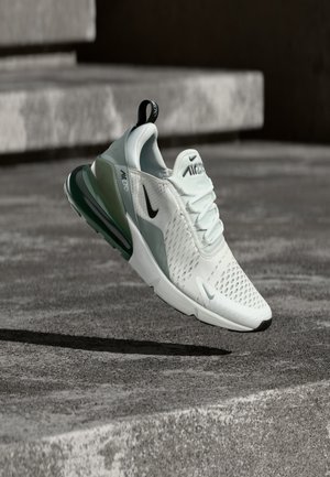 Zapatilla Nike Air Max blanca y gris suspendida sobre escalones de concreto, destacando su amortiguación y diseño transpirable.