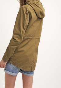 Khaki lehká parka s kapucí a volným střihem, nastavitelnou kapucí a prošívanými detaily, doplněná džínovými šortkami.