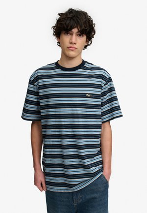 Joven con cabello rizado y oscuro, vestido con una camiseta de manga corta a rayas azules, negras y blancas, y pantalones vaqueros azules, de pie con una mano en el bolsillo.