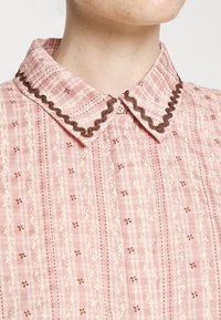 Chemise à carreaux rose avec un col marron festonné, ornée de motifs floraux et de feuilles couleur crème, et une fermeture à boutons au niveau du col.