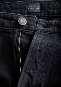Crne traperice s vidljivim zatvaračem, oznakom marke "JACK & JONES PREMIUM" na dugmetu i teksturiranim materijalom sa suptilnim uzorcima.