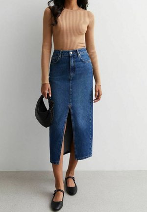 Langer Jeansrock mit einem Schlitz vorne, mittlerer blauer Waschung, kombiniert mit einem taillierten, beige-ribbed Langarmoberteil und einer schwarzen Handtasche. Flache schwarze Schuhe.