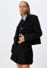 Zwarte cropped jas met gouden knopen en twee borstzakken, gedragen met een zwarte geplooide rok met hoge taille. Lichtblauwe blouse met kraag eronder.