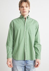 Πράσινο πουκάμισο με ρίγες, με λευκές κάθετες ρίγες, διαθέτει επισημασμένο γιακά και τσέπη στο στήθος. Κατασκευασμένο από ελαφρύ ύφασμα.