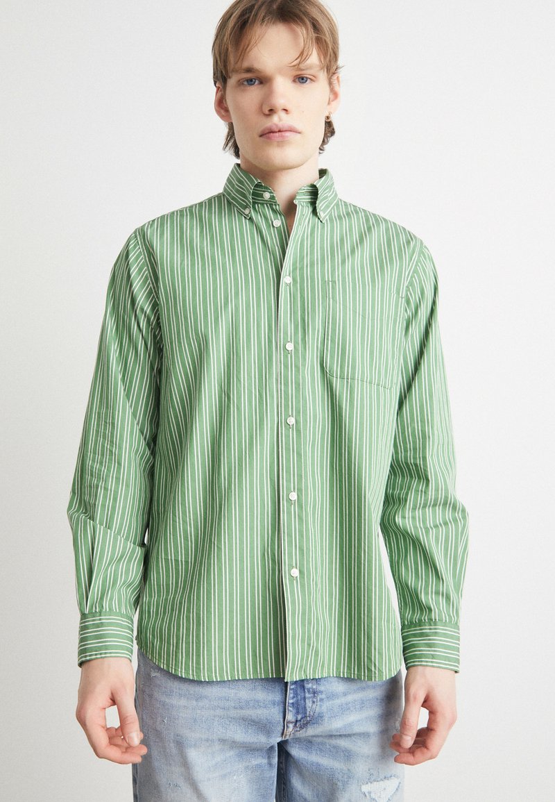 Πράσινο πουκάμισο με ρίγες, με λευκές κάθετες ρίγ�ες, διαθέτει επισημασμένο γιακά και τσέπη στο στήθος. Κατασκευασμένο από ελαφρύ ύφασμα.