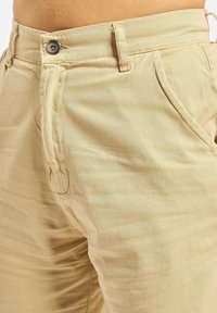 Beige katoenen broek met een rechte snit, voorzien van een metalen knoopsluiting, zijzakken en zichtbare stikseldetails.