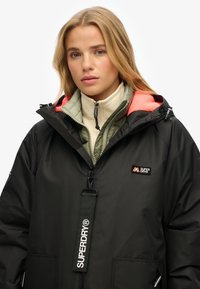 Mujer joven con chaquetas superpuestas: forro polar beige, acolchada verde y abrigo negro de Superdry con forro de capucha naranja.