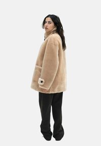 Beige Kunstfelljacke mit hohem Kragen, Seitentasche und kontrastierenden Knopfdetails am Ärmel. Lockerer Schnitt, kombiniert mit schwarzen Hosen.