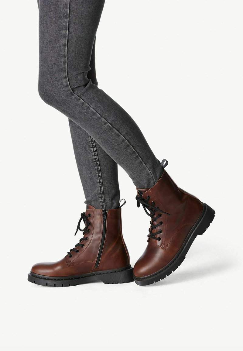 Stivali alla caviglia in pelle marrone con lacci davanti, cerniera laterale, suola in gomma nera e cuciture a contrasto. Abbinati a jeans slim-fit grigi.