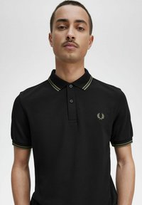 Hombre con una camiseta polo negra con finas rayas color oliva en el cuello y las mangas, y un pequeño logo de corona de laurel en el pecho izquierdo.