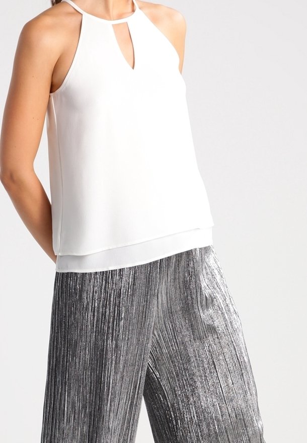 Haut blanc sans manches à col halter avec ouverture devant, associé à un pantalon large plissé métallisé argenté sur une personne.