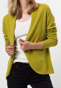 Gelber Cardigan mit Strukturmuster über einem weißen Hemd, mit offenem, lockerem Vorderteil und langen Ärmeln, kombiniert mit einer schwarzen Hose.