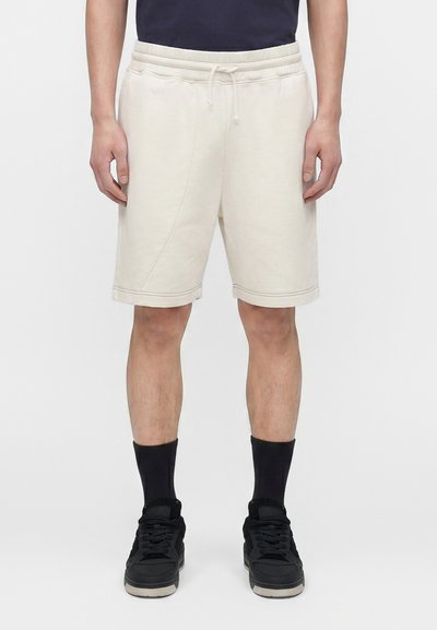 Homme portant un short crème avec cordon, des chaussettes noires montantes et des baskets noires, debout devant un fond blanc uni.