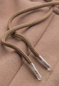 Hnědé pletené šnůrky na texturované tkanině, s průhlednými plastovými špičkami s raženým logem. Měkké a ležérní designové prvky.