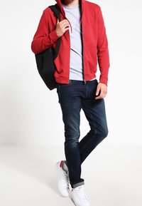 Sudadera roja con cremallera, camiseta gris debajo, jeans azules y zapatillas blancas. Mochila negra colgada del hombro. Diseño simple.