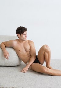 Schwarze Schwimmbriefe, getragen von einem Model mit durchtrainierter Figur, das auf einer weichen, strukturierten, hellen Fläche vor einem schlichten Hintergrund sitzt.