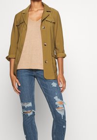 Giacca utilitaria verde oliva con maniche arrotolate, caratterizzata da bottoni argento e tasche a pattina, abbinata a una maglia beige a scollo a V e jeans blu strappati.