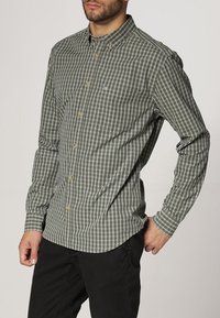 Mężczyzna w zielono-szarej koszuli w kratę z kołnierzykiem i czarnych spodniach, stojący z jedną ręką na biodrze.