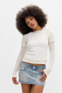 Ung kvinna med stor afrofrisyr som bär lager av vita toppar med långa ärmar och en kort denimkjol, poserar mot en vit bakgrund.