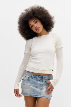 Jeune femme avec une grande coiffure afro, portant des hauts blancs superposés à manches longues et une jupe en denim courte, posant sur fond blanc.