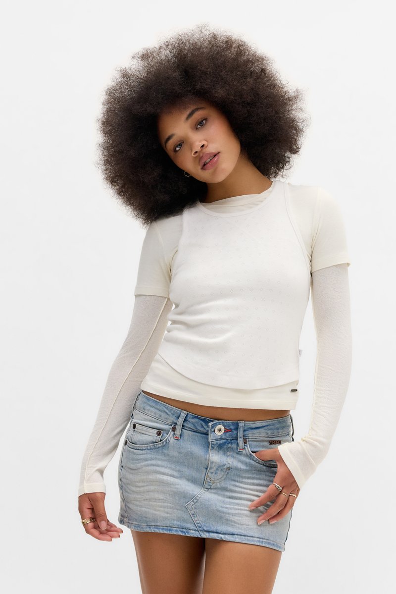 Ung kvinna med stor afrofrisyr som bär lager av vita toppar med långa ärmar och en kort denimkjol, poserar mot en vit bakgrund.
