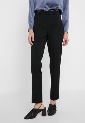 Frau trägt schwarze, schmal zulaufende Hose, blaues Langarmtop und schwarze Slingback-Pumps mit Absatz vor einfachem weißem Hintergrund.