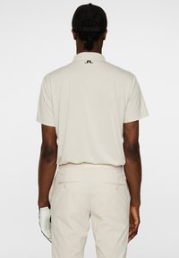 Beige polo-t-shirt med korta ärmar, avslappnad passform och en diskret logotyp på baksidan. Matchas med ljusa byxor och en handske.