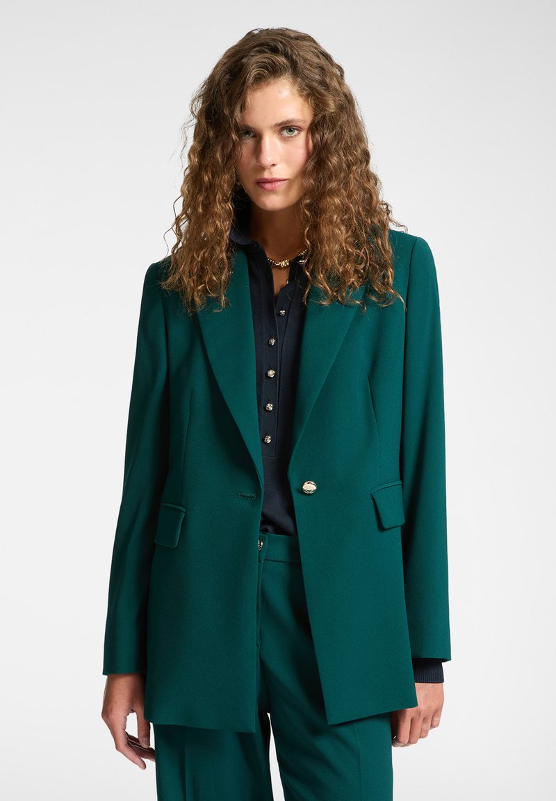 Blazer verde con revers affilati, due tasche frontali e un'unica chiusura con bottone dorato, abbinato a una camicia scura sotto.