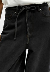 Pantalones de denim negros con un alto talle, que incluyen un cinturón atado, costuras en contraste naranja y bolsillos frontales. Textura suave, corte relajado.