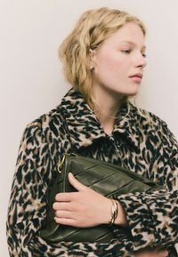 Blondýnka v kabátu s leopardím vzorem drží prošívanou olivově zelenou kabelku na jednobarevném pozadí.