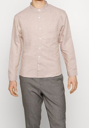 Homme portant une chemise beige à boutons avec col officier et un pantalon gris, debout devant un fond blanc uni.